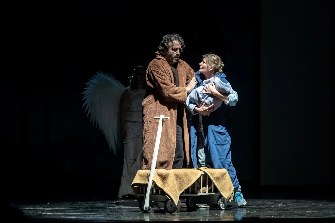 Zwei Menschen stehen auf der Bühne und halten ein Baby. Eine Person mit Flügeln steht hinter ihnen. Die Bühne ist dunkel.