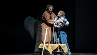 Zwei Menschen stehen auf der Bühne und halten ein Baby. Eine Person mit Flügeln steht hinter ihnen. Die Bühne ist dunkel.