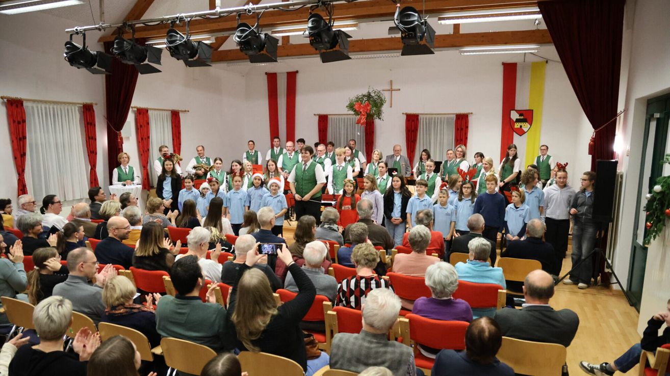 Ein Chor tritt auf der Bühne in einem Saal mit roten Vorhängen und einer Kreuzdekoration auf. Zuschauer sitzen in roten Stühlen und klatschen. Ein Mann macht ein Foto mit einem Handy.