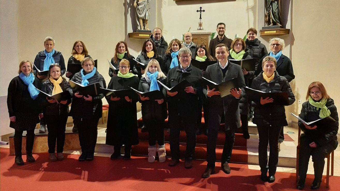 Ein Chor steht auf einem roten Teppich in einer Kirche, hält Musikbücher und hat eine Statue und ein Kreuz im Hintergrund.