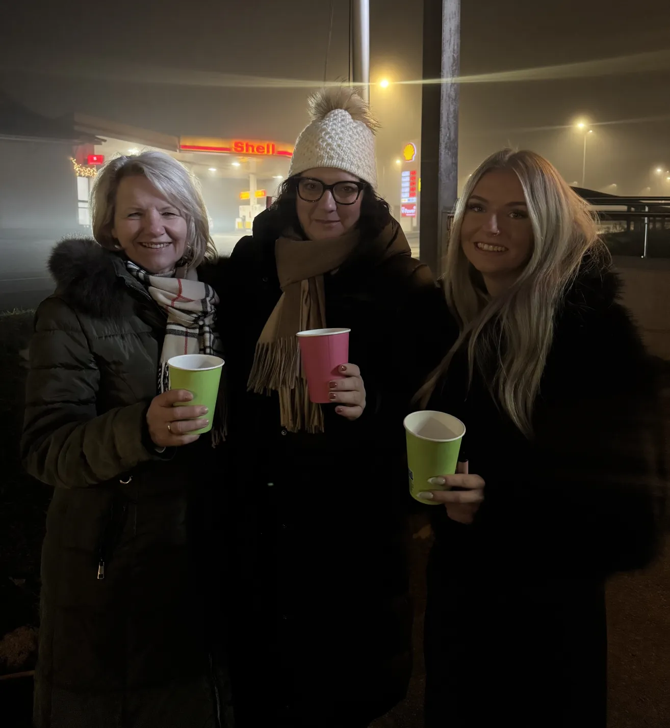 Drei Frauen stehen nebeneinander, lächeln und halten grüne Becher, einer mit einem rosa Becher. Sie sind an einer Tankstelle bei Nacht.
