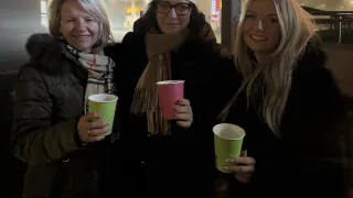 Drei Frauen stehen nebeneinander, lächeln und halten grüne Becher, einer mit einem rosa Becher. Sie sind an einer Tankstelle bei Nacht.