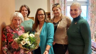 Fünf Frauen stehen lächelnd zusammen. Die Frau vorn hält einen Blumenstrauß. Die Frau in der Mitte trägt eine hellblaue Blazer. Die Frau rechts trägt ein gehäkeltes Oberteil.