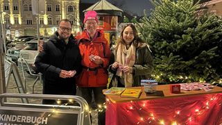 Drei Personen stehen vor einem Weihnachtsbaum und einem kleinen Tisch mit Büchern. Ein McDonald's-Schild ist im Hintergrund sichtbar.