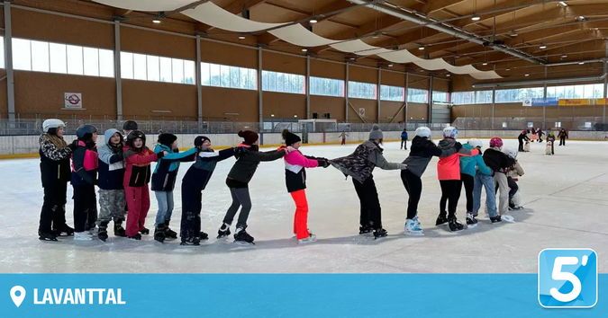 Eine Gruppe von Menschen skatet auf einer großen Indoor-Eisbahn. Sie halten sich an den Händen und bilden einen Kreis. Sie tragen Winterkleidung und Helme. Die Eisbahn hat große Fenster.