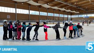 Eine Gruppe von Menschen skatet auf einer großen Indoor-Eisbahn. Sie halten sich an den Händen und bilden einen Kreis. Sie tragen Winterkleidung und Helme. Die Eisbahn hat große Fenster.