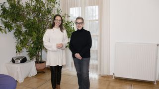 Zwei Frauen stehen vor einem Fenster, lächeln und posieren für ein Foto. Die Frau auf der linken Seite trägt einen weißen Pullover und Lederhosen. Die Frau auf der rechten Seite trägt ein schwarzes Rollkragenshirt und Jeans. Ein Topf mit einer Pflanze und ein Heizkörper sind im Hintergrund zu sehen.