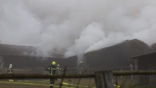 Feuerwehrleute in voller Ausrüstung bekämpfen ein Feuer in einem Scheunengebäude, aus dem starker Rauch aufsteigt. Die Szene ist durch Nebel verschleiert.
