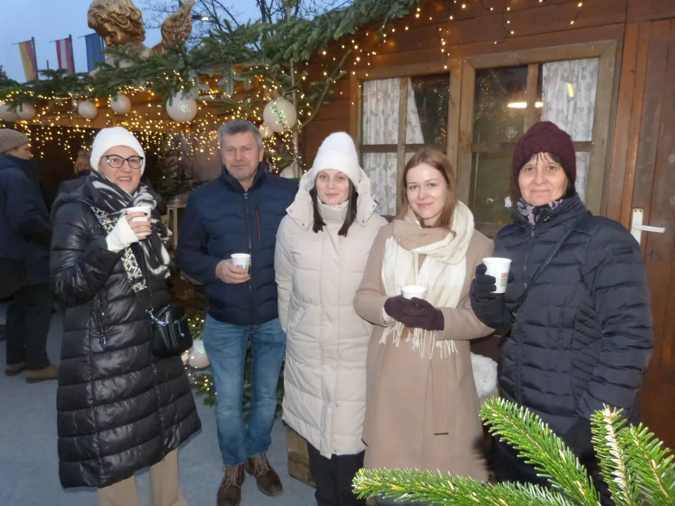 Fünf Personen posieren für ein Foto vor einer festlich geschmückten Struktur, verziert mit Weihnachtslichtern und Ornamenten. Sie halten alle Tassen in den Händen und scheinen die festliche Atmosphäre zu genießen.