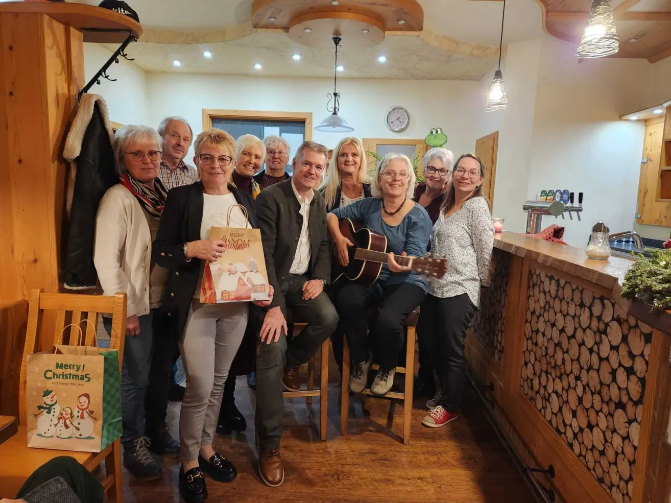 Eine Gruppe von Menschen versammelt sich in einem gemütlichen Raum, einige sitzen auf Hockern, andere stehen. Ein Mann hält eine Gitarre, und eine Frau hält einen Papierbeutel. Die Umgebung umfasst eine Holztheke, Regale und eine Froschuhr.