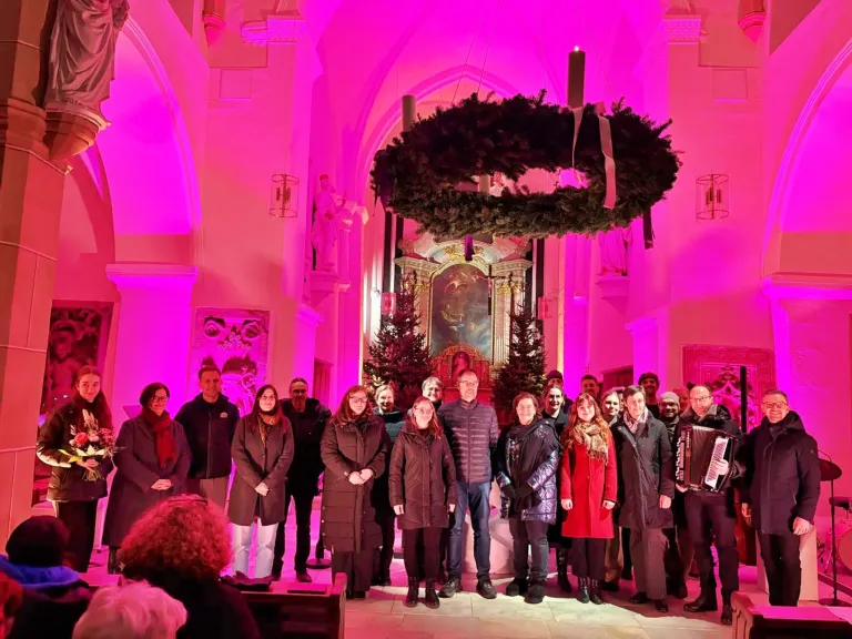 Eine Gruppe von Menschen posiert für ein Foto in einer Kirche mit einem Kranz und Weihnachtsbäumen. Rosa Lichter erhellen den Raum.