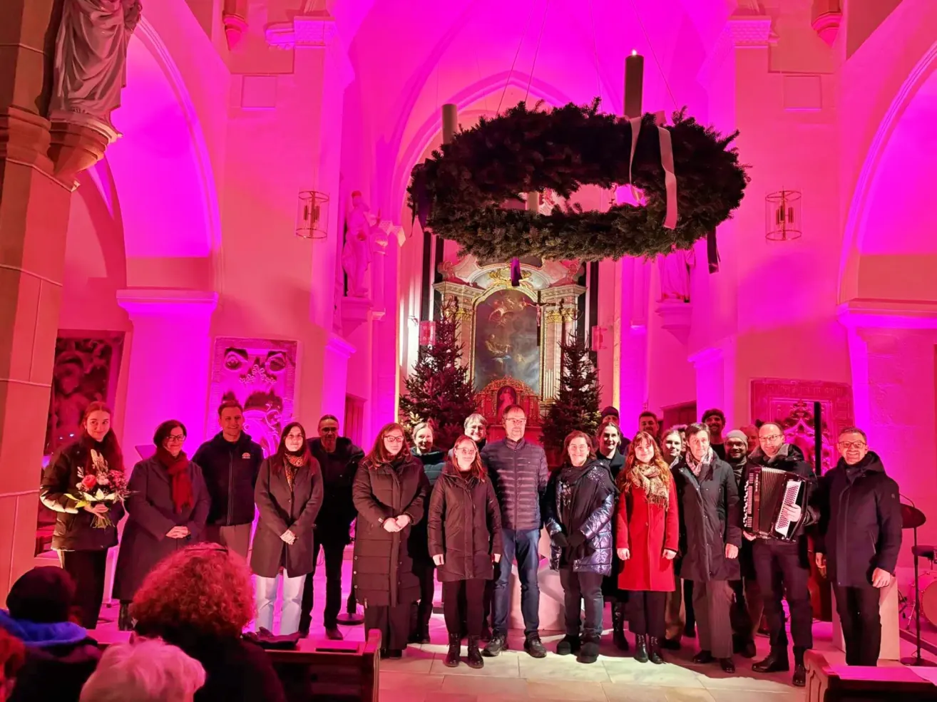 Eine Gruppe von Menschen posiert für ein Foto in einer Kirche mit einem Kranz und Weihnachtsbäumen. Rosa Lichter erhellen den Raum.