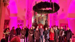 Eine Gruppe von Menschen posiert für ein Foto in einer Kirche mit einem Kranz und Weihnachtsbäumen. Rosa Lichter erhellen den Raum.