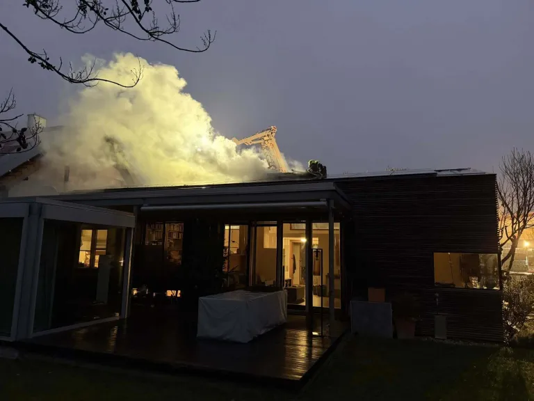 Ein Feuer brennt auf dem Dach eines modernen Hauses in der Dämmerung, mit Feuerwehrleuten auf dem Dach. Rauch steigt vom Dach auf, und ein Tisch mit einer Abdeckung steht auf der Terrasse.