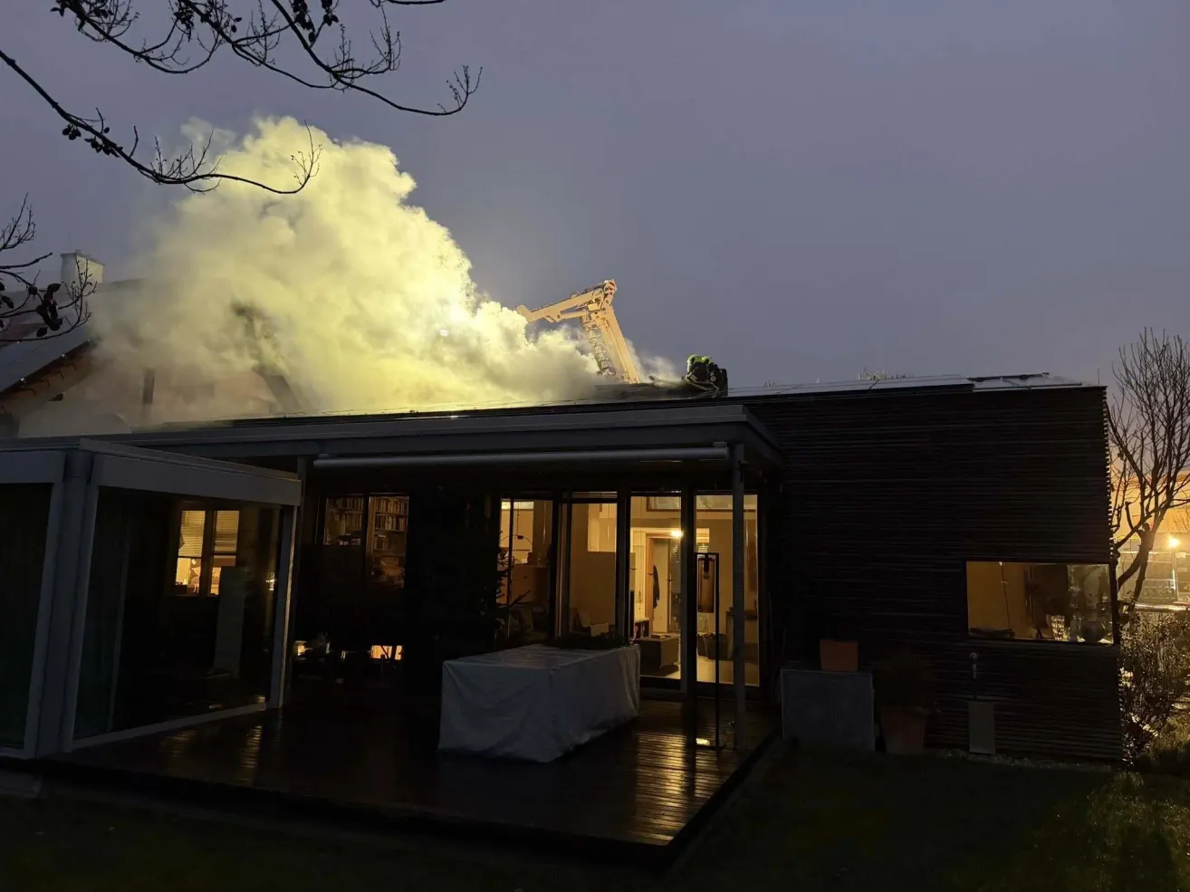 Ein Feuer brennt auf dem Dach eines modernen Hauses in der Dämmerung, mit Feuerwehrleuten auf dem Dach. Rauch steigt vom Dach auf, und ein Tisch mit einer Abdeckung steht auf der Terrasse.
