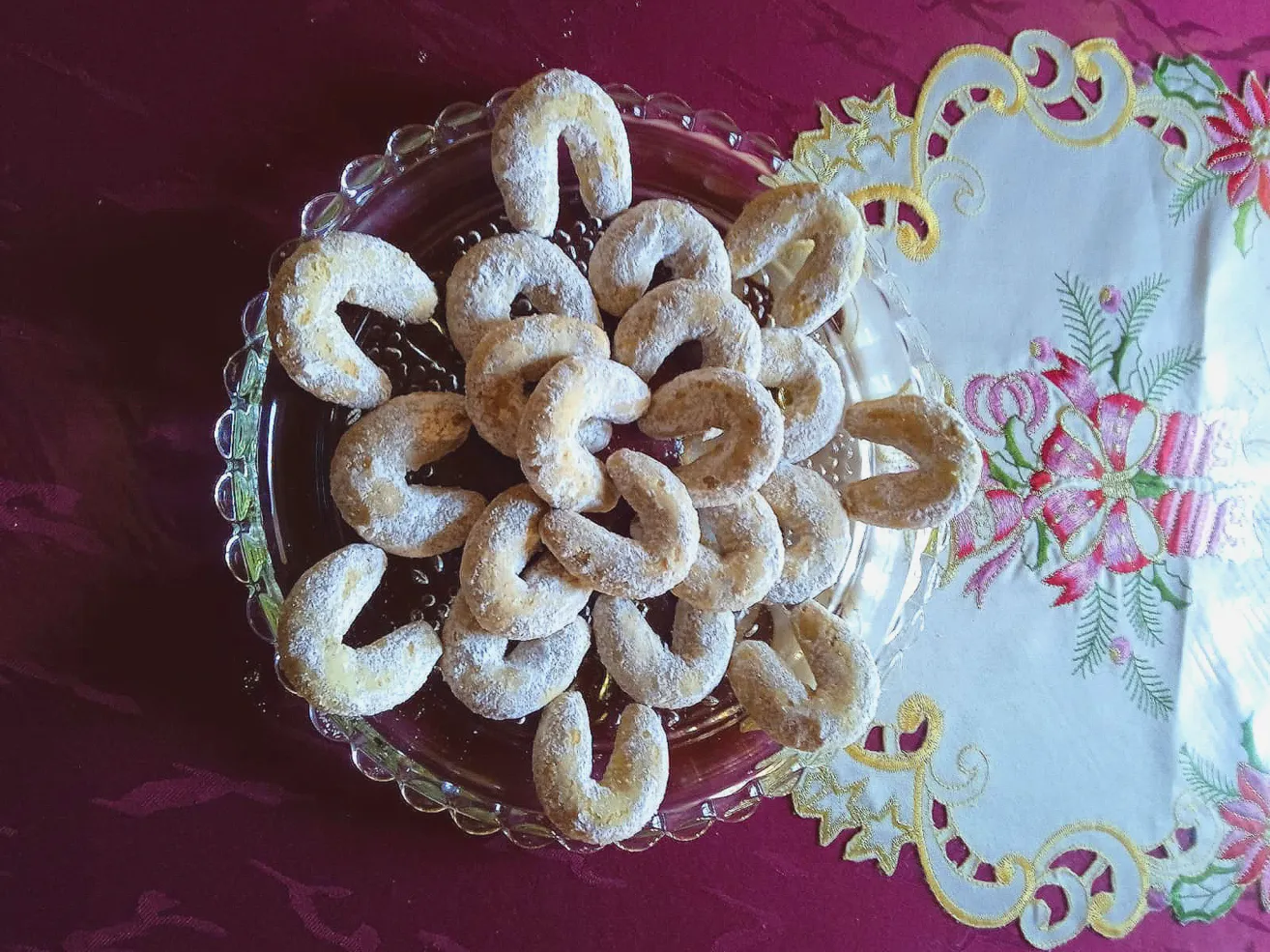 Eine Glasschale mit halbmondförmigen Gebäckstücken, bestreut mit Puderzucker, steht auf einem purpurroten Tischtuch mit einer blumigen Spitze.
