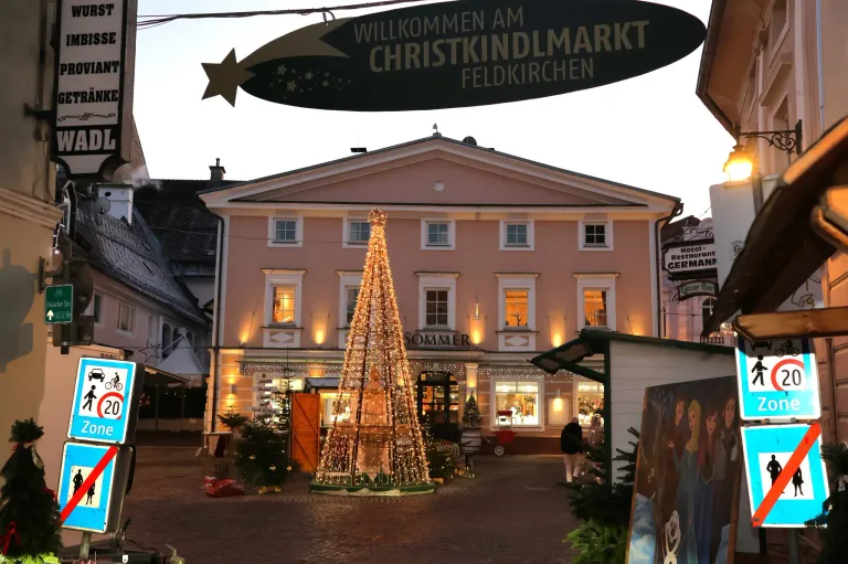 Ein geschmückter Weihnachtsbaum steht vor einem rosa Gebäude mit einem Schild, das 'Willkommen am Christkindlmarkt Feldkirchen' besagt. Das Gebäude ist beleuchtet und hat mehrere Fenster. Eine Person geht auf der rechten Seite.