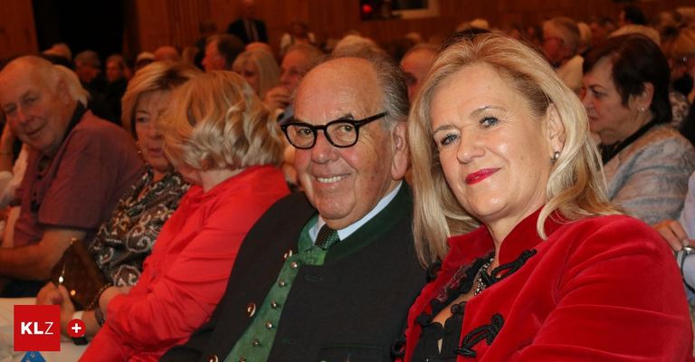 Ein älteres Paar lächelt in einem Theater in die Kamera. Der Mann trägt eine Brille und eine grüne Weste. Die Frau trägt einen roten Mantel mit schwarzer Spitze. Dahinter sitzt ein Publikum.