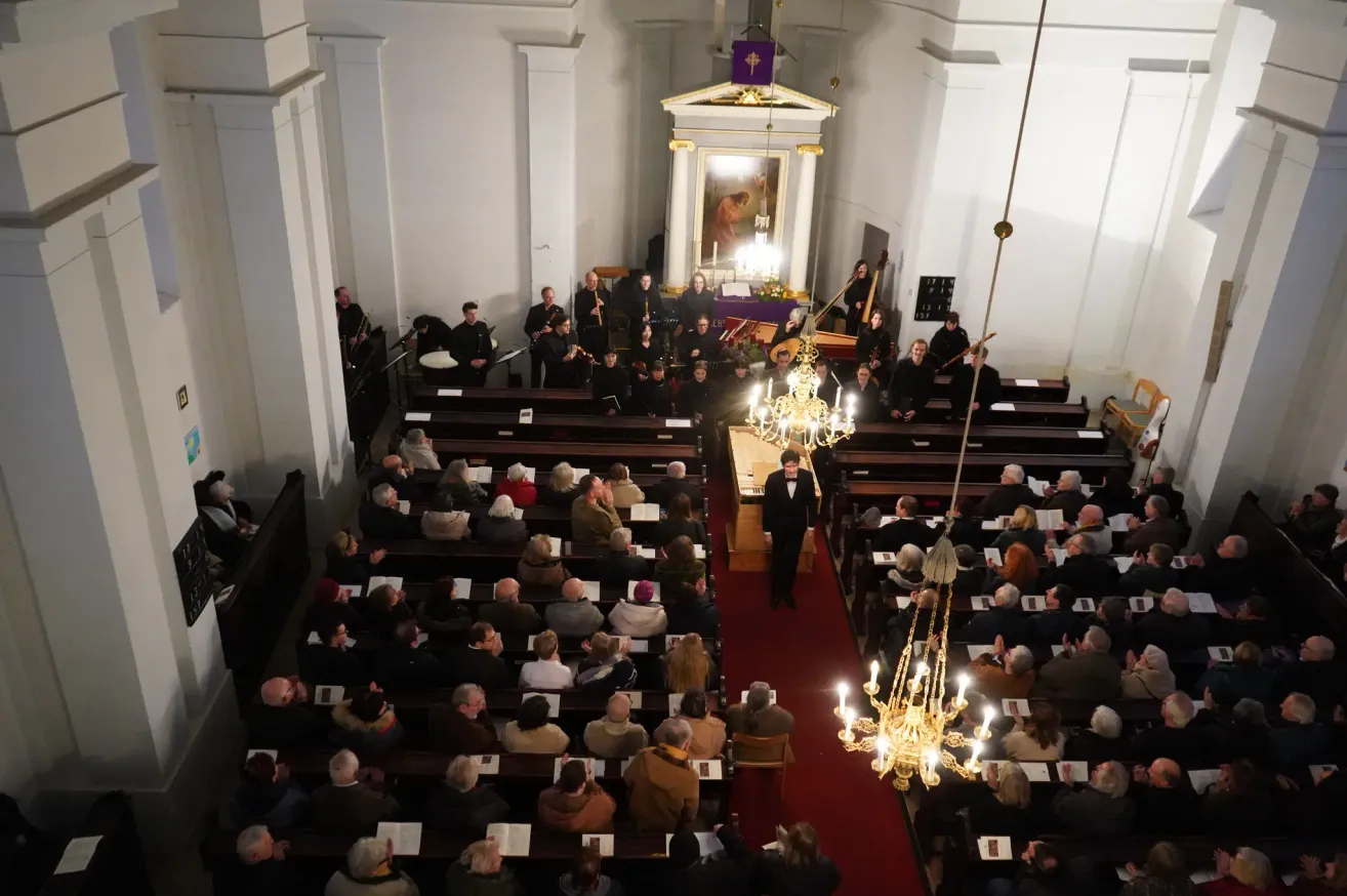 A church filled with people. A choir is performing at the altar. The audience is sitting in rows. A conductor is leading the choir. The church has a red carpet, chandeliers, and a cross on the altar.