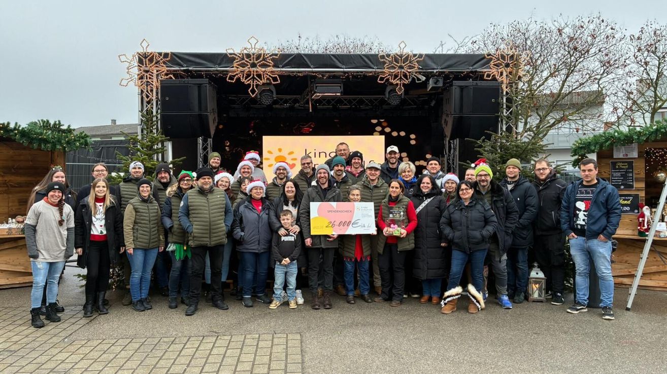 Eine Gruppe von Menschen posiert für ein Foto vor einer Bühne mit einem Banner. Sie tragen alle Winterkleidung und einige tragen Mützen. Ein Mann in der Mitte hält einen Scheck und eine Trophäe. Hinter ihnen steht ein Weihnachtsbaum und eine dekorierte Bühne mit Lichtern und Lautsprechern.