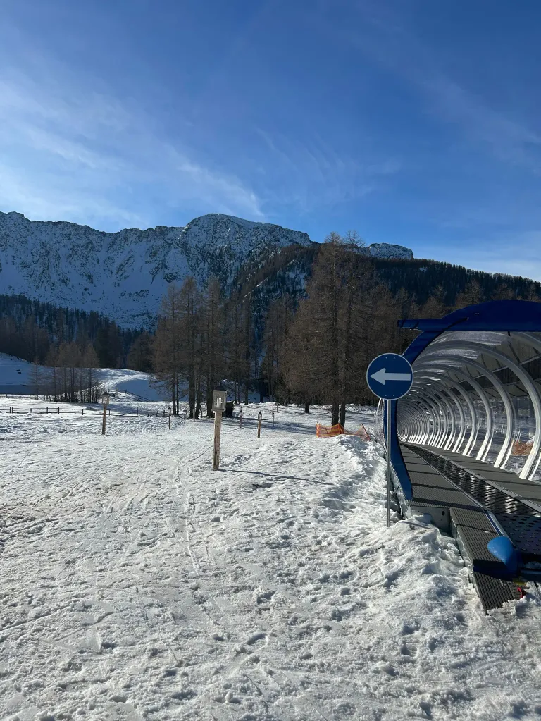 Schneebedeckte Landschaft mit Bergen im Hintergrund. Ein blauer Himmel mit Wolken. Bäume umgeben einen verschneiten Hang mit einem Skilift.