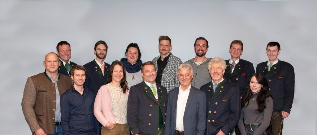 Eine Gruppe von Erwachsenen in formeller Kleidung, einschließlich Männer und Frauen, posiert für ein Foto. Einige tragen Krawatten und Jacken. Sie lächeln und stehen vor einer weißen Wand.