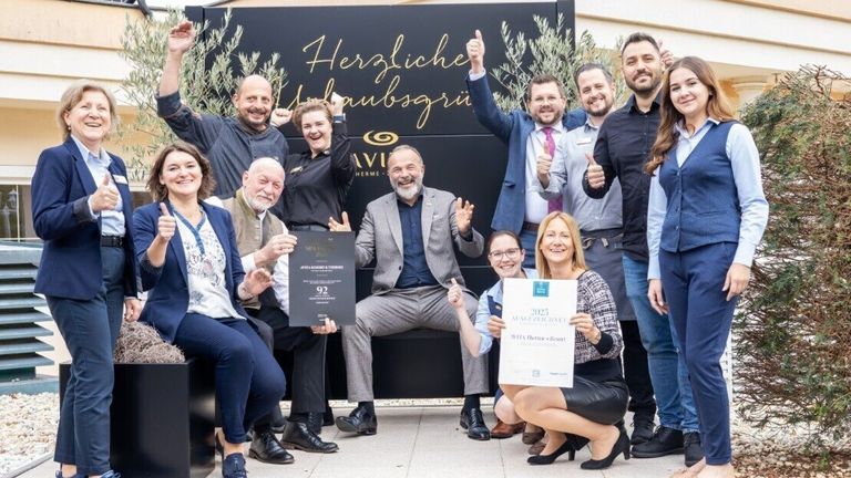 Eine Gruppe von Menschen steht lächelnd zusammen und hält Auszeichnungen. Hinter ihnen steht ein schwarzes Schild mit goldener Schrift, das 'Herzliche Willkommen' besagt. Sie scheinen zu feiern.