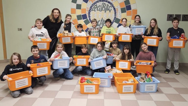 Eine Gruppe von Kindern und Erwachsenen posiert für ein Foto, während sie Spendenboxen mit der Aufschrift 'Bewegung Mitmensch Spendebox' halten. Sie lächeln und stehen auf einem gemusterten Boden.