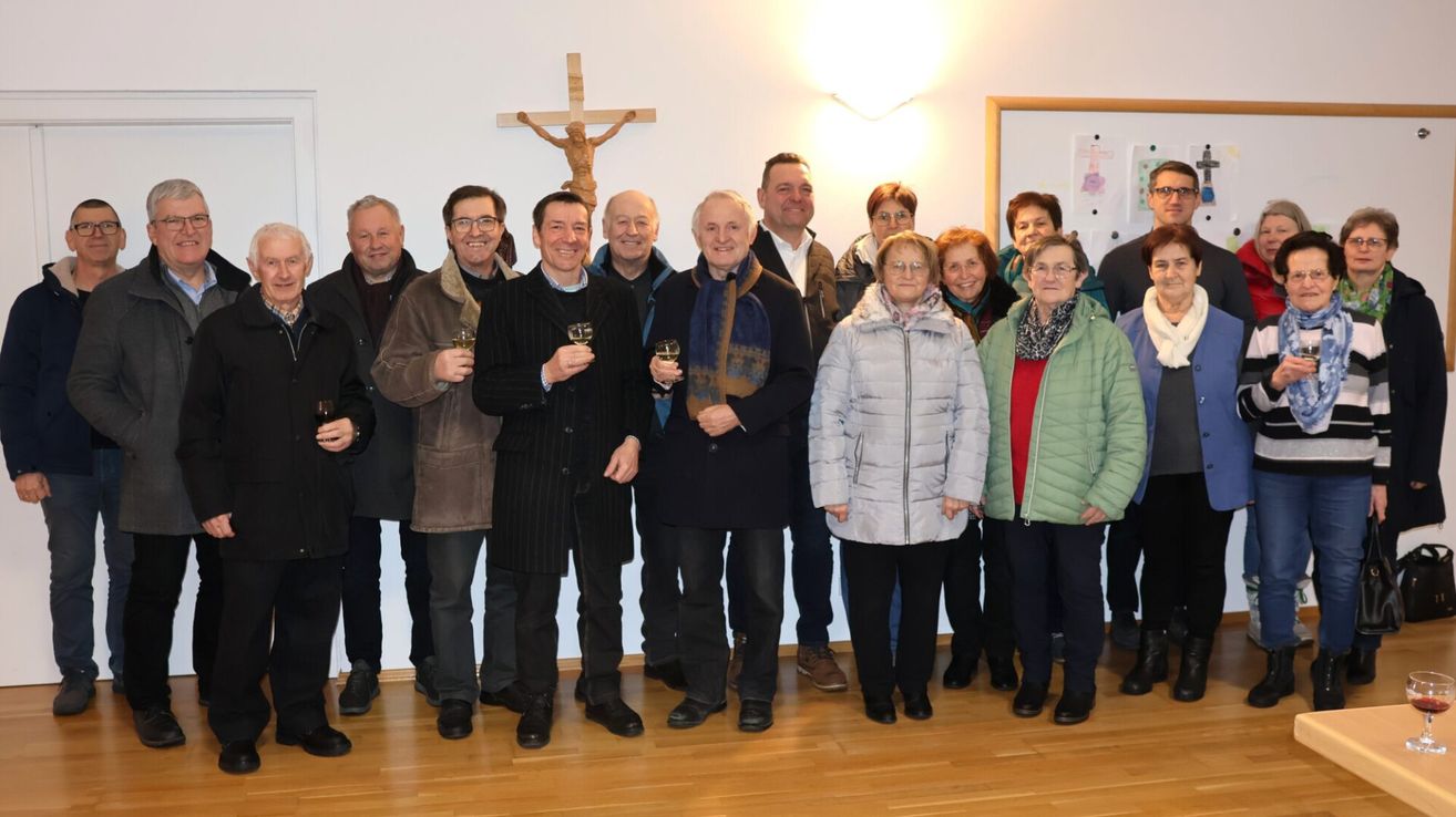 Eine Gruppe von Menschen steht in einem Raum mit einem Kreuz an der Wand. Sie alle lächeln und halten Gläser mit Wein in den Händen.