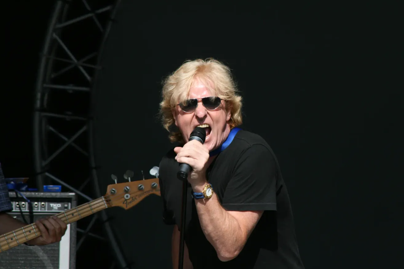 Ein Mann mit blonden Haaren, der eine Sonnenbrille und ein schwarzes T-Shirt trägt, singt in ein Mikrofon auf der Bühne. Im Hintergrund ist eine Gitarre mit schwarzem und silbernem Korpus zu sehen.