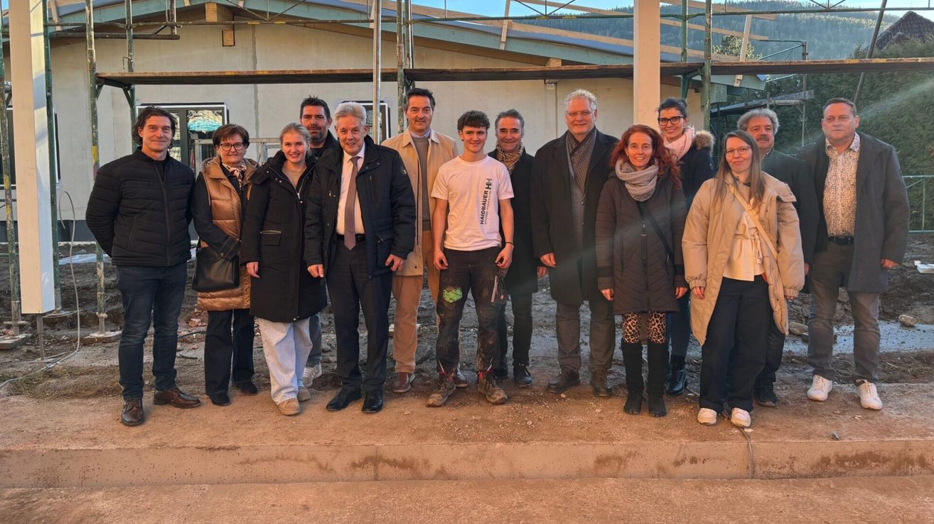 Eine Gruppe von Menschen in Winterkleidung, einige mit Brille, steht vor einer Baustelle und lächelt für die Kamera.
