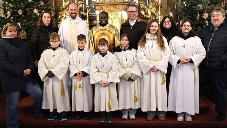 Eine Gruppe von Kindern in weißen Gewändern mit gelben Schärpen steht vor einem Priester in einer Kirche und posiert für ein Foto.