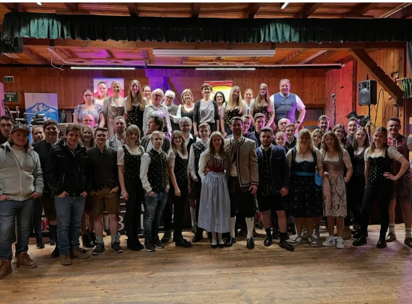 Eine große Gruppe von Menschen in traditioneller Kleidung steht auf einer Holzbühne, einige lächeln und andere posieren für ein Foto, während ein Mikrofon im Hintergrund aufgestellt ist.