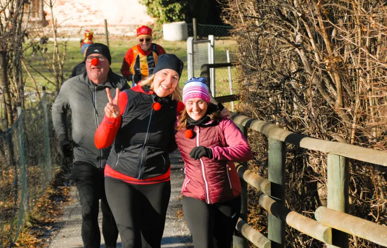 Zwei Frauen und ein Mann mit Clownsnase laufen im Freien. Sie tragen Winterkleidung und stehen neben einem Holzzaun.