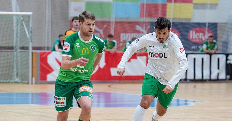 Zwei Männer spielen Futsal, einer in Grün und der andere in Weiß, laufen aufeinander zu mit konzentrierten Gesichtsausdrücken. Der Hintergrund zeigt ein verschwommenes Publikum und einen roten Banner mit weißem Text.