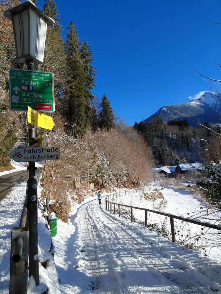 Eine Person geht auf einem verschneiten Pfad mit einem Wegweiser, der auf Mondsee und St. Wolfgang hinweist. Der Pfad ist von einem Zaun gesäumt, und es gibt Bäume und Berge im Hintergrund.