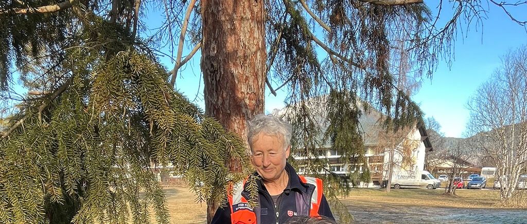 Eine ältere Frau steht lächelnd unter einem Baum auf einem Feld und trägt eine reflektierende Sicherheitsweste. Hinter ihr ist ein weißer Lieferwagen vor einem Gebäude mit vielen Fenstern geparkt. Der Himmel ist klar und blau.
