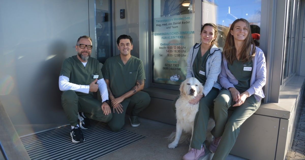 Vier Personen und ein Hund posieren vor einer Tierarztpraxis. Das Schild sagt Tiermedizinisches Zentrum für Kleintiere. Es ist Montag bis Freitag von 9 Uhr bis 12 Uhr und 14 Uhr bis 18 Uhr geöffnet. Die Telefonnummer ist 02167 7303.