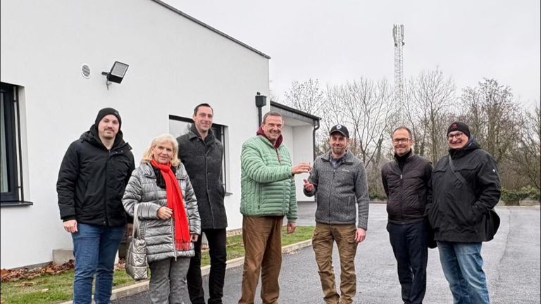 Fünf Personen stehen vor einem Gebäude und lächeln. Ein Mann überreicht einem anderen ein Dokument. Sie tragen Winterkleidung, und im Hintergrund sind ein weißes Gebäude, Bäume und ein Turm zu sehen.