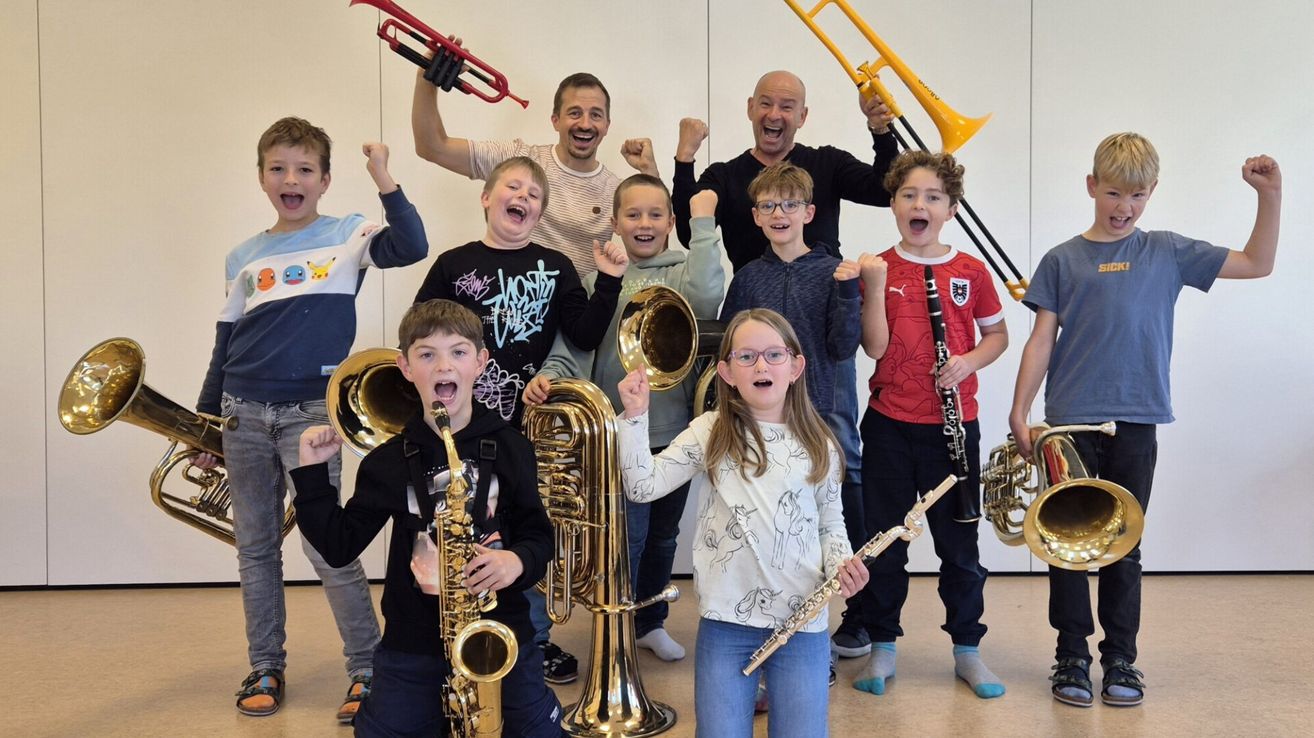 Eine Gruppe von Kindern und zwei Erwachsenen posiert für ein Foto mit Musikinstrumenten. Die Kinder lächeln alle und scheinen Spaß zu haben. Sie halten eine Trompete, ein Tenorhorn und eine Saxophon. Die Erwachsenen stehen hinter ihnen und einer von ihnen hält eine Trompete.