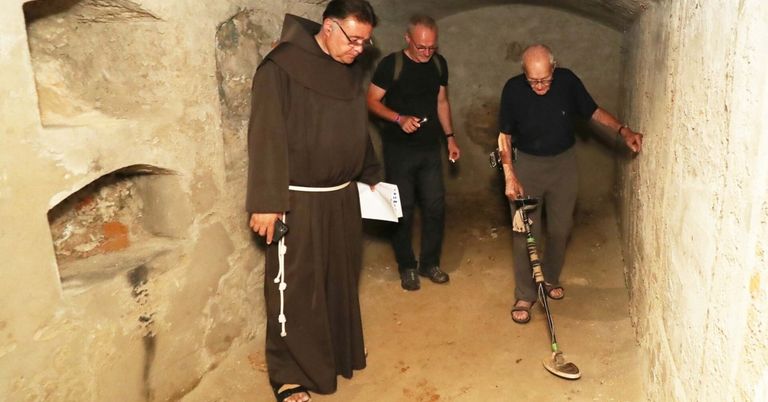 Drei Männer in einem Tunnel, einer trägt eine Mönchskutte, die anderen beiden haben Metalldetektoren. Der Mann mit der Brille schaut auf sein Handy.