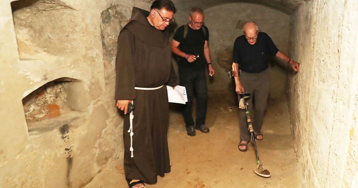 Drei Männer in einem Tunnel, einer trägt eine Mönchskutte, die anderen beiden haben Metalldetektoren. Der Mann mit der Brille schaut auf sein Handy.