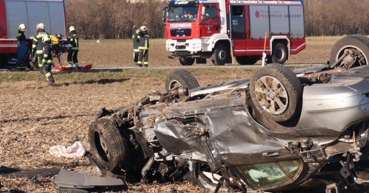 Ein umgekipptes Auto liegt auf der Seite auf einer Straße, ein Feuerwehrwagen in der Nähe. Zwei Feuerwehrleute stehen hinter einer Barriere und beurteilen wahrscheinlich die Situation.