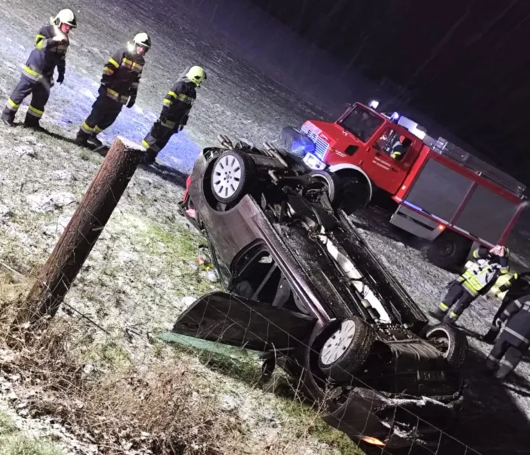 Drei Feuerwehrleute inspizieren ein umgekipptes Auto mit einem roten Feuerwehrwagen im Hintergrund bei Nacht.