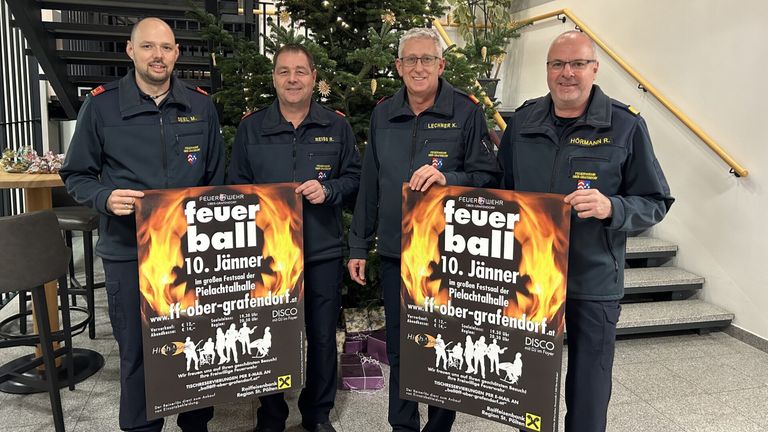 Vier Feuerwehrmänner in Uniform halten Plakate für die Feuerball-Veranstaltung am 10. Januar hoch. Die Veranstaltung findet in der Pielachtalhalle in Ober-Grafendorf statt. Die Plakate haben ein feueriges Design und enthalten Veranstaltungsdetails.