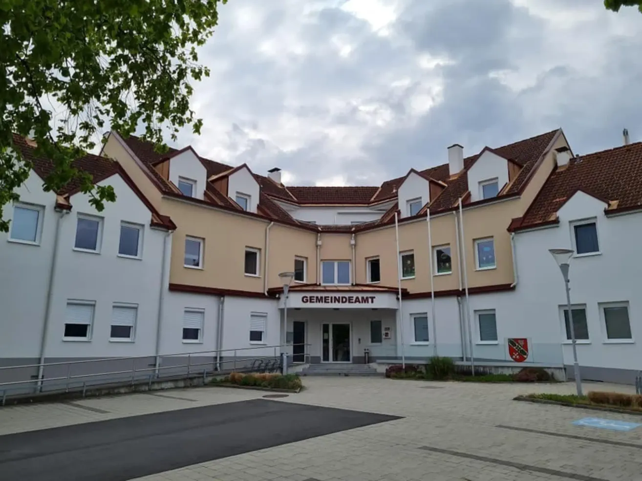 Ein großes weißes Gebäude mit braunen Dächern und Fenstern hat das Wort 'Gemeindeamt' darauf geschrieben. Es gibt Flaggen an Pfosten in der Nähe des Eingangs.