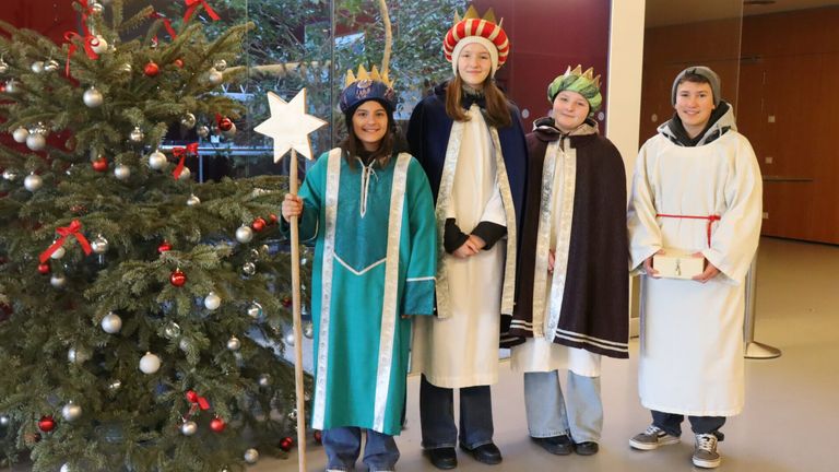 Vier Personen in Kostümen stehen vor einem Weihnachtsbaum. Die Frau auf der linken Seite hält einen Sternenstab, während die anderen Kronen tragen. Sie scheinen Teil einer festlichen Feier zu sein.