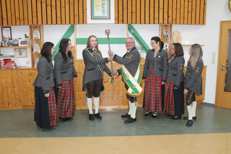 Ein Mann in traditioneller Kleidung übergibt einer Frau einen Stab, während andere in Uniform um sie herum in einem Raum mit Holzwänden und Regalen stehen.