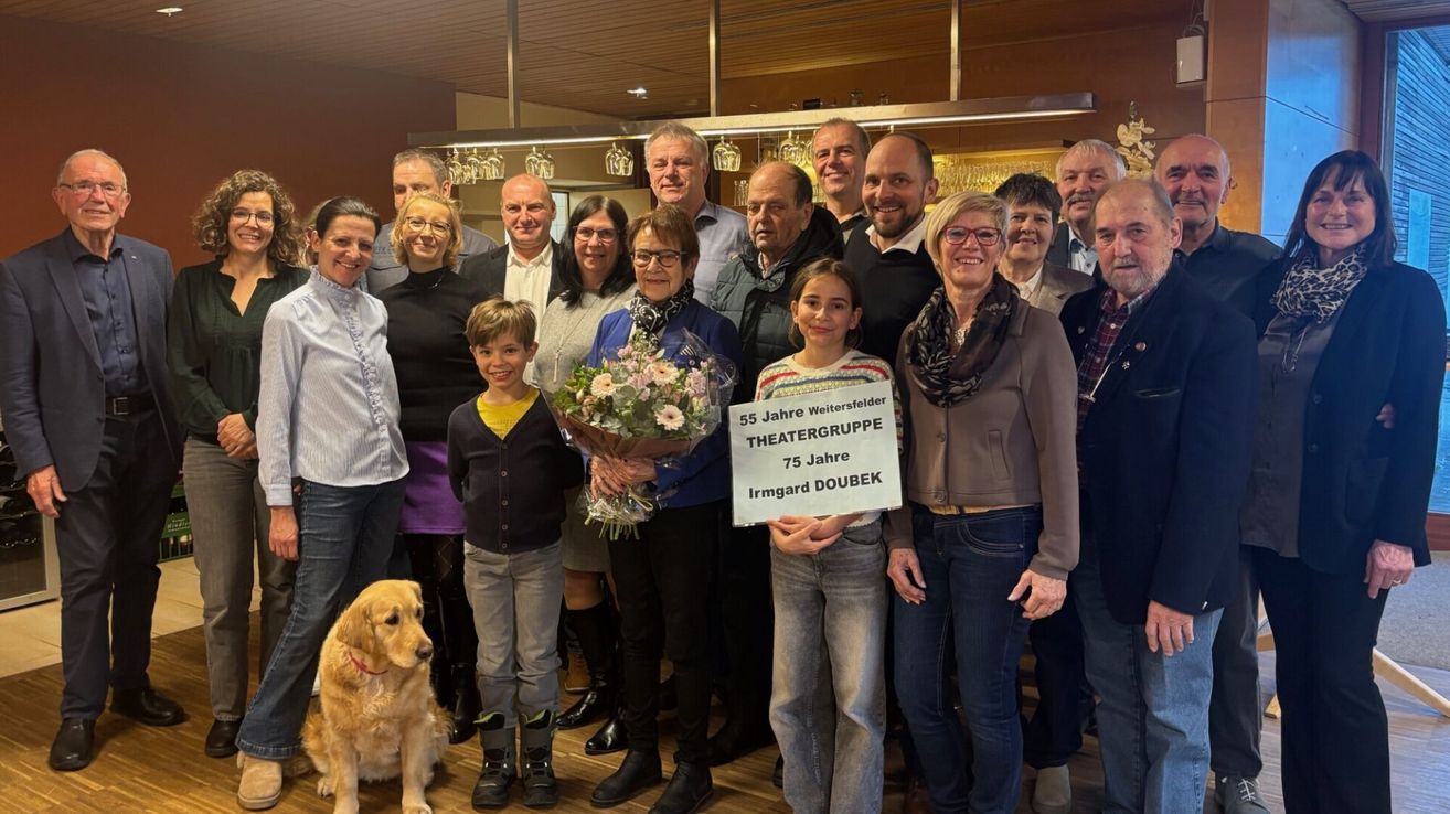 Eine Gruppe von Menschen steht in einem Raum zusammen und feiert einen Meilenstein. Eine Frau hält ein Schild und Blumen, und ein Kind steht neben ihr. Das Schild sagt '55 Jahre Wettersfelder Theatergruppe' und '75 Jahre Irmgard Doubek'.