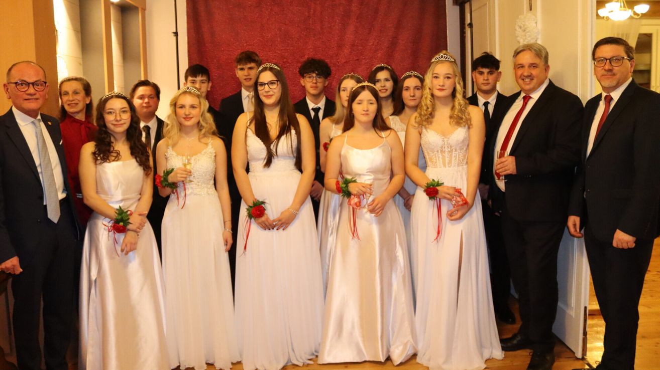 Eine Gruppe junger Frauen in weißen Kleidern und Blumenkränzen steht zusammen, lächelt und posiert für ein Foto. Sie halten Weingläser und einige haben rote Blumen an ihren Kleidern. Dahinter ist ein roter Vorhang und einige Männer in Anzügen zu sehen.